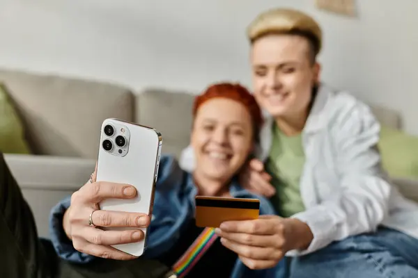 A lesbian couple with short hair smiles while taking a selfie together using a credit card at home. — Stock Photo