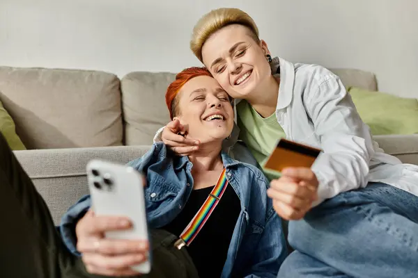 Two women, a lesbian couple, sit on a couch with a credit card in hand, making a purchase together. — Stock Photo