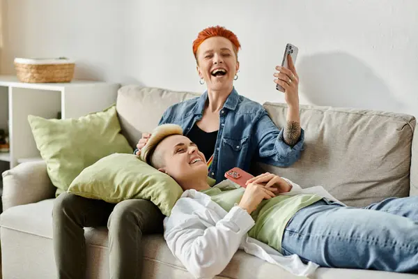 Dos mujeres, una pareja de lesbianas con el pelo corto, se sientan de cerca en un sofá, absortos en su teléfono celular. - foto de stock