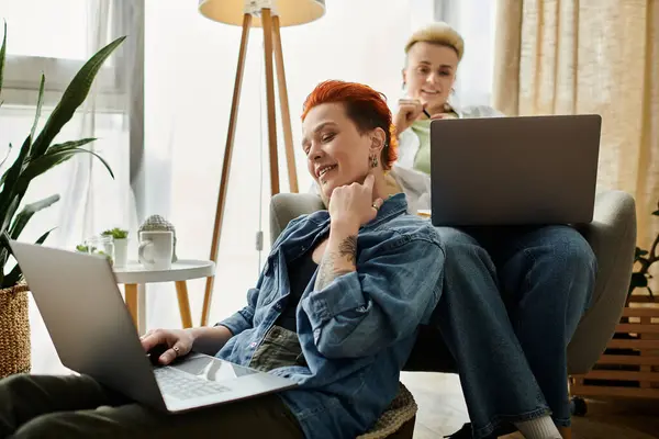 Couple lesbien aux cheveux courts assis sur une chaise, utilisant des ordinateurs portables pour travailler ou naviguer sur Internet à la maison. — Photo de stock