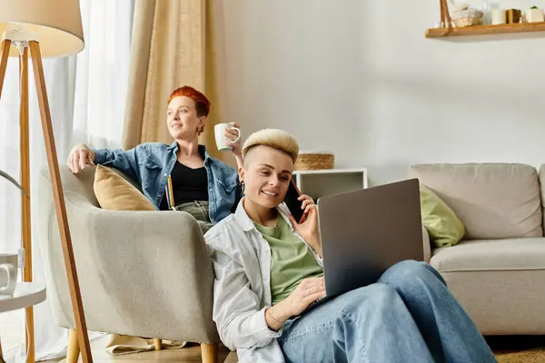 Um casal lésbico relaxa em um sofá, tomando café e usando um laptop juntos. — Fotografia de Stock