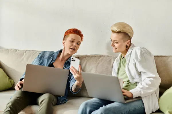Two women, a lesbian couple with short hair, sit on a couch engrossed in their laptops at home. — Stock Photo