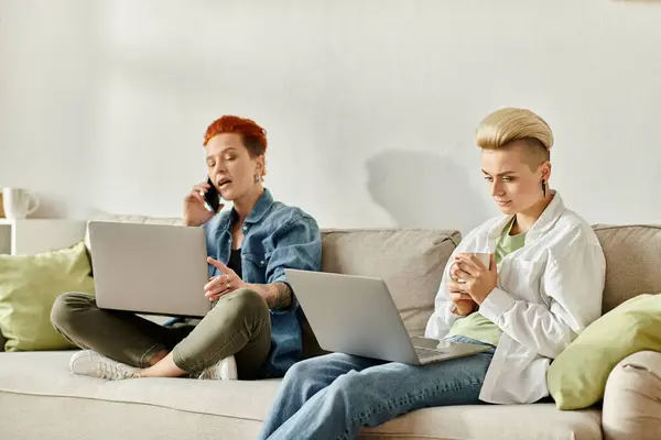 Two women with short hair sit side by side on a couch, each absorbed in their own laptop screen. — Stock Photo
