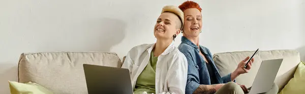 Lesbian couple with short hair engrossed in work on laptops while sitting closely on a couch at home. — Stock Photo