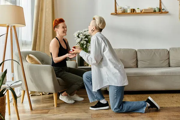 One woman kneels, offering flowers and ring to another in a cozy living room setting. — Stock Photo