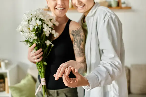 Dos mujeres sostienen ramos de flores en una acogedora sala de estar, mostrando anillo de compromiso - foto de stock