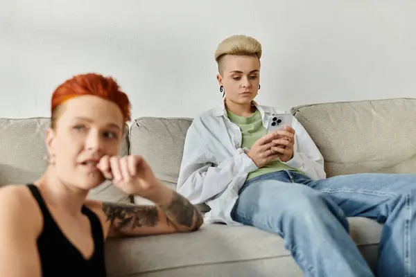 Zwei Personen, ein lesbisches Paar mit kurzen Haaren, sitzen auf einer Couch, die in ein Telefon vertieft ist, getrennt voneinander. — Stockfoto