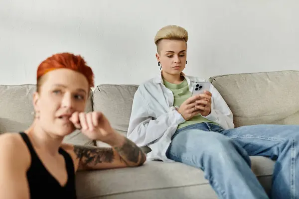 Zwei Personen, ein lesbisches Paar mit kurzen Haaren, sitzen auf einer Couch, die in ein Telefon vertieft ist, getrennt voneinander. — Stockfoto