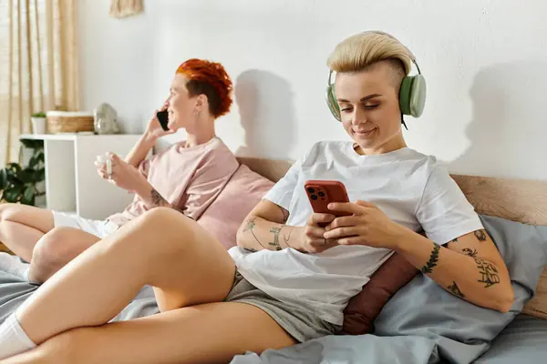 Two people, a lesbian couple, sitting on a bed and listening to music with headphones on in their bedroom. — Stock Photo