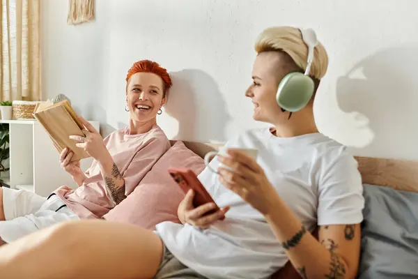 A lesbian couple with short hair sitting together on a bed, engrossed in reading a book, showcasing their bond and shared interests. — Stock Photo
