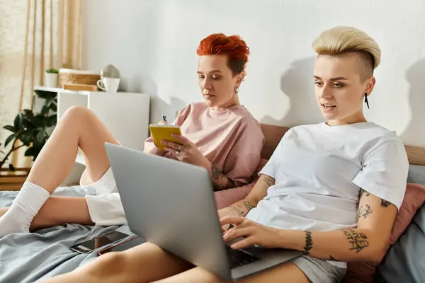 Deux femmes, un couple de lesbiennes aux cheveux courts, assises sur un lit ensemble, absorbées par l'utilisation d'un ordinateur portable. — Photo de stock