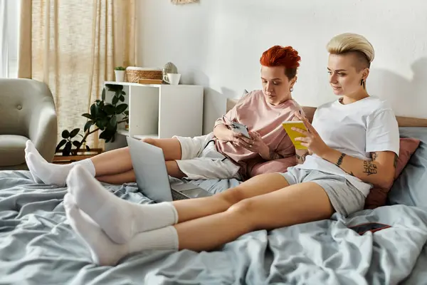 A lesbian couple with short hair sitting on a bed, engaged in using a laptop together. — Stock Photo