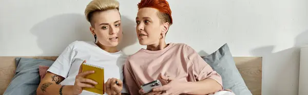 A lesbian couple with short hair sitting closely on a bed, holding a cell phone, engaged in a warm and intimate moment. — Stock Photo