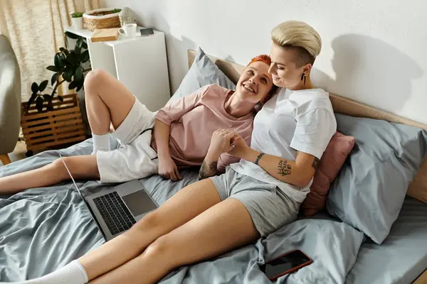 Дві жінки з коротким волоссям сидять на ліжку, занурені у використання ноутбука разом у затишній обстановці спальні. — стокове фото