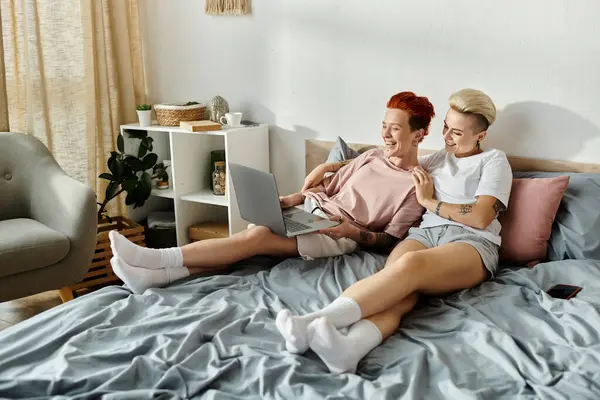 Two women with short hair sit on a bed, engrossed in using a laptop. — Stock Photo