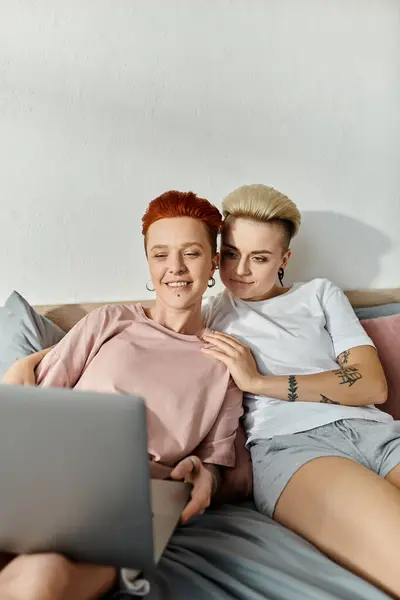 Deux femmes, un couple de lesbiennes, s'assoient ensemble sur un lit, absorbées dans leur ordinateur portable, partageant un moment dans leur chambre confortable. — Photo de stock