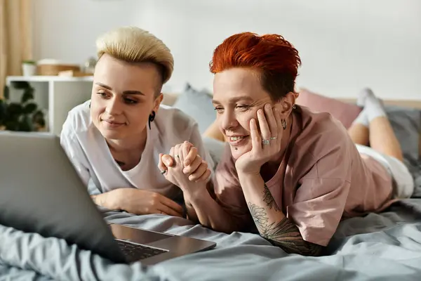 Двоє людей, лесбійська пара з коротким волоссям, розслабляються і з'єднуються на ліжку, дивлячись на екран ноутбука. — стокове фото