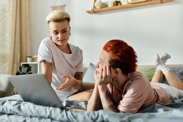 Two people, a lesbian couple, sit on a bed with short hair, focused on a laptop screen in a cozy bedroom. — Stock Photo