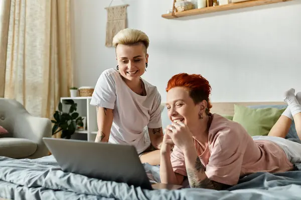 Two women with short hair lounging on a bed, using a laptop together in a bedroom, embracing a cozy moment. — Stock Photo