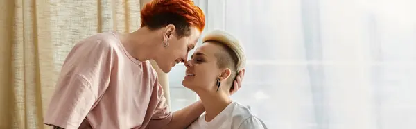 Two women with short hair sharing a tender kiss in front of a window in a cozy bedroom, celebrating their love in an intimate moment. — Stock Photo