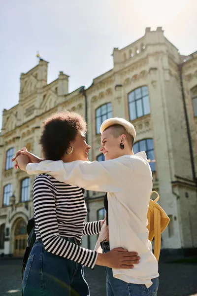 Deux jeunes femmes d'ethnies différentes s'embrassent chaleureusement devant une toile de fond architecturale époustouflante, symbolisant le lien et l'amitié. — Photo de stock