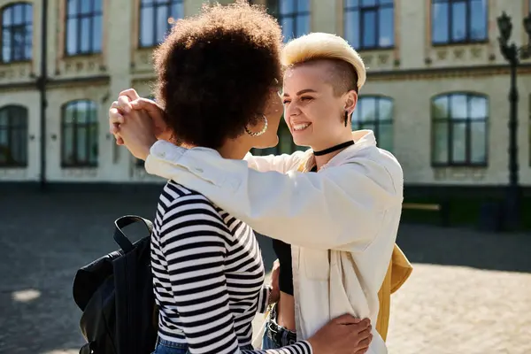 Two young women from diverse backgrounds embrace warmly in front of a historic building, showcasing love — Stock Photo