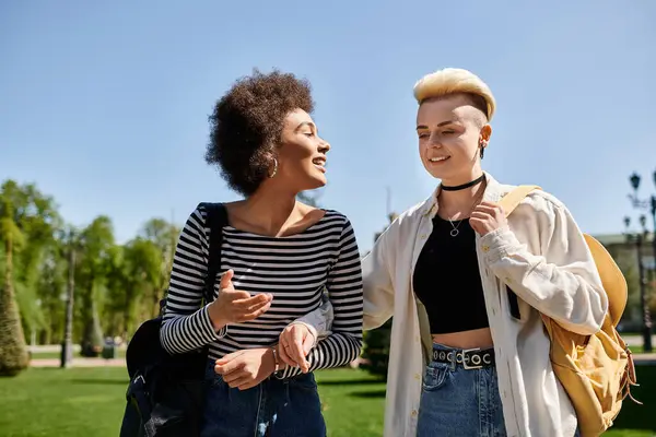 Dos mujeres jóvenes, una pareja lésbica multicultural, vestidas con estilo charlando en un parque cerca de un campus universitario. - foto de stock