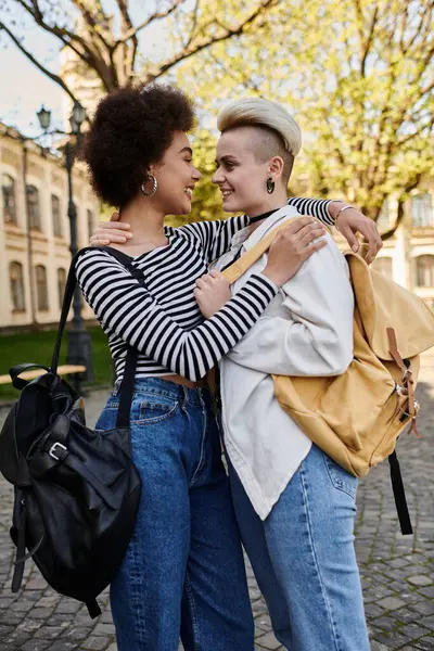 Due giovani donne, una nera e una caucasica, si abbracciano calorosamente mentre tengono gli zaini all'aperto. — Foto stock