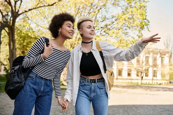 Two young women, a multicultural lesbian couple, hold hands as they walk down a street near a university campus. — Stock Photo
