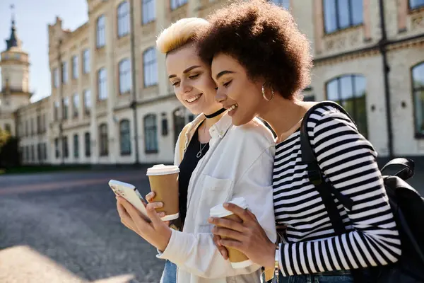 Zwei junge Frauen in stylischer Kleidung, in ein Handy vertieft, stehen vor einem modernen Gebäude an einer belebten Stadtstraße. — Stockfoto