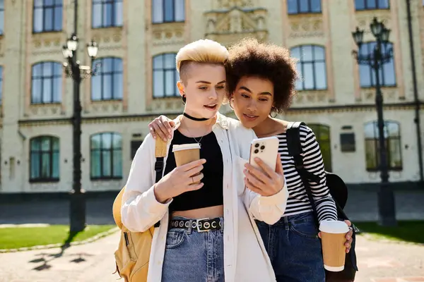 Due giovani donne in abbigliamento casual, assorbite nel loro cellulare, si trovano di fronte a un edificio moderno su una vivace strada cittadina. — Foto stock