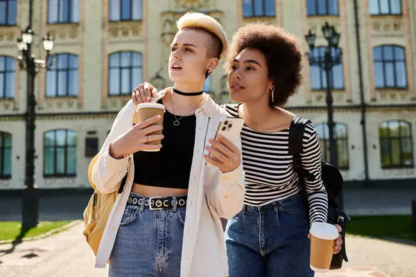 Deux jeunes femmes, un couple lesbienne multiculturel, se promènent dans une rue près d'une université avec des tasses à café à la main. — Photo de stock