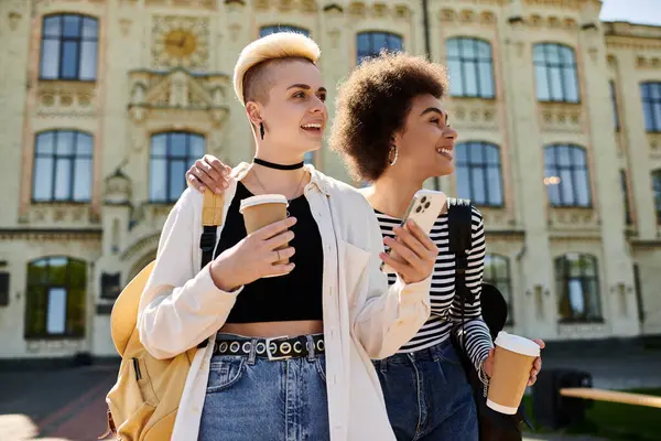 Two young women, a multicultural lesbian couple, stroll leisurely down the street, each holding a coffee cup. — Stock Photo