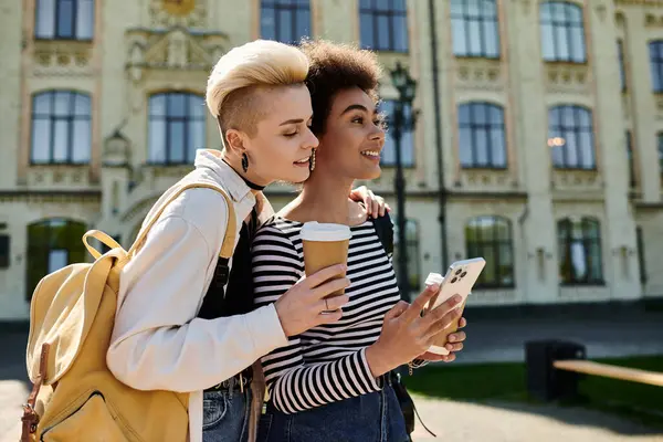 Two young women in casual attire standing in front of a building, engrossed in phone — Stock Photo