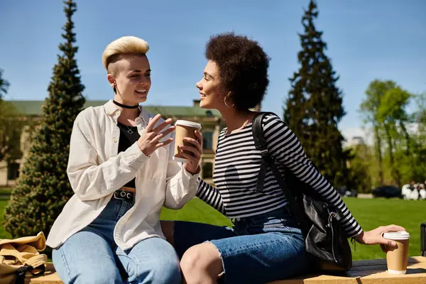 A multicultural lesbian couple, stylishly dressed, sit and chat happily on a bench in a park on a sunny day. — Stock Photo