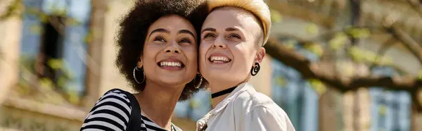 Two young women, a multicultural lesbian couple, share joyful smiles in front of a university building. — Stock Photo