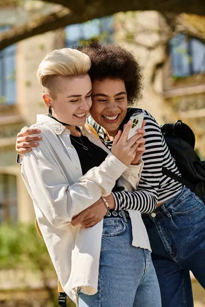 Zwei junge Frauen in lässiger Kleidung umarmen sich, während sie in ihre Handys vertieft sind, und scheinen einen besonderen Moment miteinander zu teilen. — Stockfoto