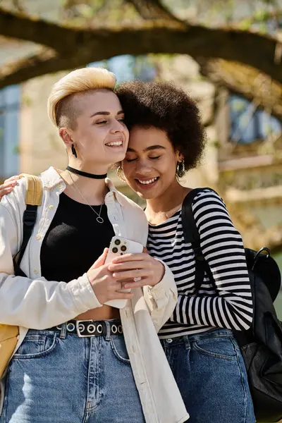 Due giovani donne si abbracciano mentre sono assorte nel telefono, condividendo un momento di connessione tra le distrazioni della tecnologia. — Foto stock