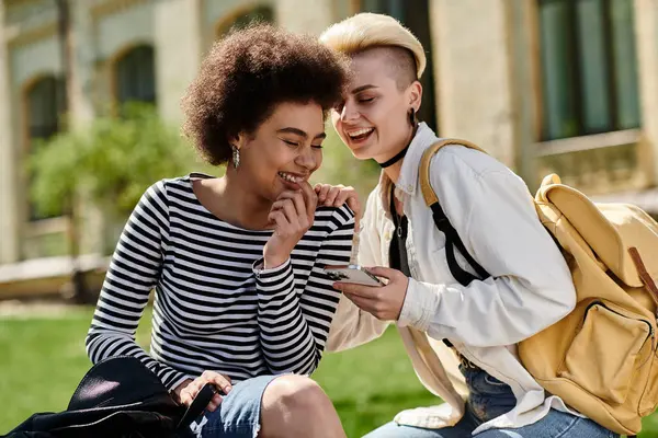 Two young girls in stylish attire sitting on the grass, engrossed in phone, amidst a serene setting. — Stock Photo