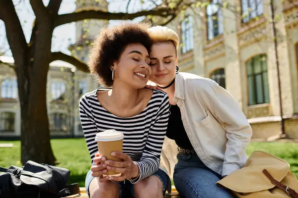 Two young women in stylish attire sit on a bench, sipping coffee and engaged in conversation outdoors. — Stock Photo