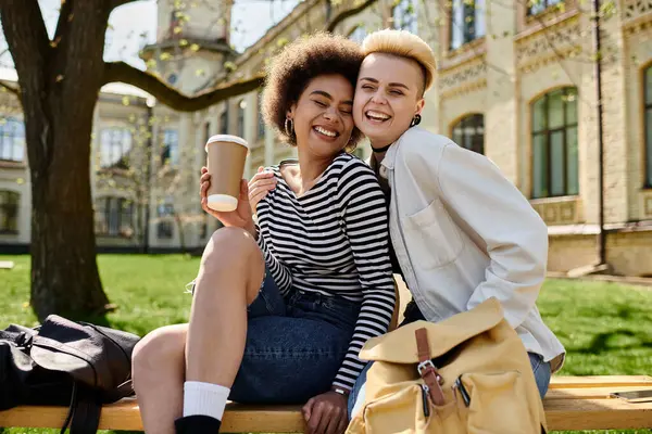Due giovani ragazze con abbigliamento elegante siedono su una panchina, sorseggiando caffè in un momento sereno all'aperto. — Foto stock