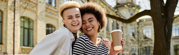 Young, multicultural lesbian couple in stylish attire walk down street holding coffee cups near university campus. — Stock Photo