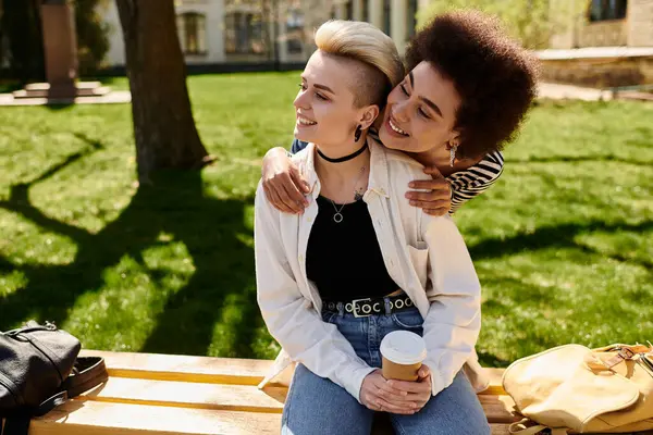 Due giovani ragazze, una afroamericana e una caucasica, si abbracciano affettuosamente seduti su una rustica panchina di legno. — Foto stock