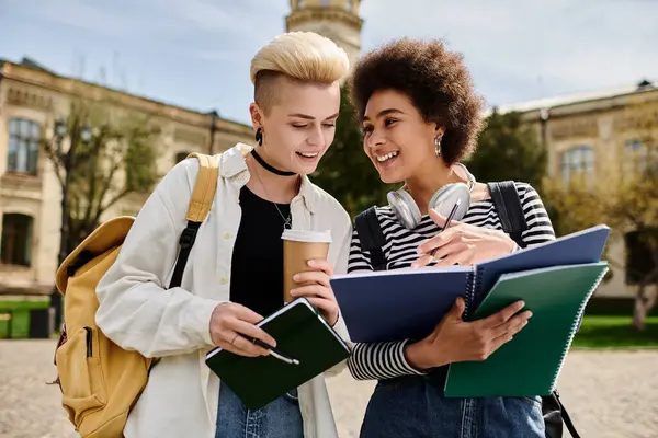 Ein multikulturelles lesbisches Paar spricht, während es Notizbücher vor einem Gebäude auf einem Universitätscampus hält. — Stockfoto