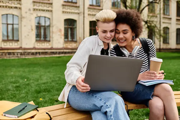 Due donne in abbigliamento casual sedute su una panchina, concentrate su uno schermo portatile, in un ambiente urbano. — Foto stock