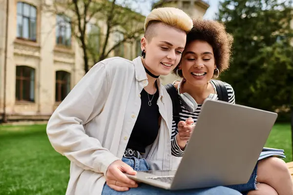 A stylish lesbian couple sitting on a bench, browsing a laptop together near a university campus. — Stock Photo