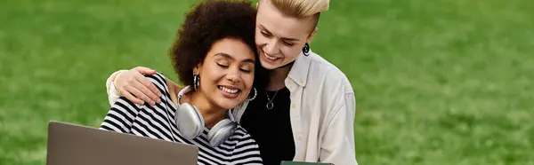 Two women in casual clothing hugging each other while typing on a laptop outdoors near a university campus. — Stock Photo