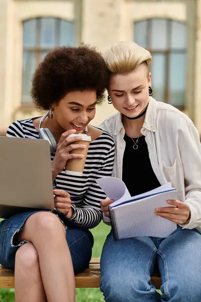 Two young girls in casual attire sitting on a bench, working on a laptop and sipping coffee in a cozy outdoor setting. — Stock Photo