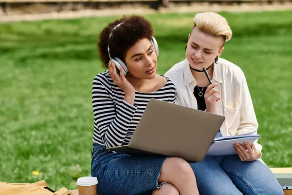 Duas mulheres jovens, elegantemente vestidas, sentadas na grama usando um laptop. — Fotografia de Stock