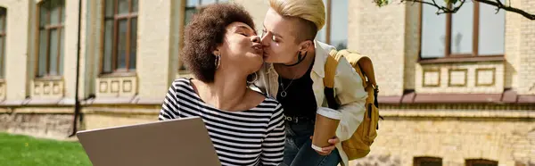 Deux jeunes femmes partagent un tendre baiser tout en tenant un ordinateur portable devant un bâtiment animé sur le campus. — Photo de stock